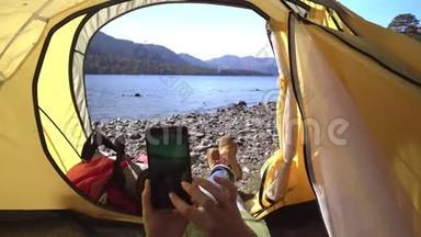 女旅行者躺在湖边的一个黄色帐篷里，手里拿着智能手机，然后是